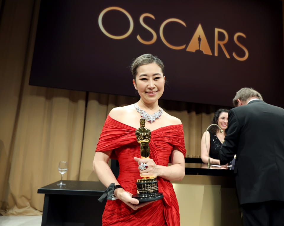Sheridan graduate Maggie Kang is pictured in a red dress holding her Academy Award trophy with a black Oscars banner in the background