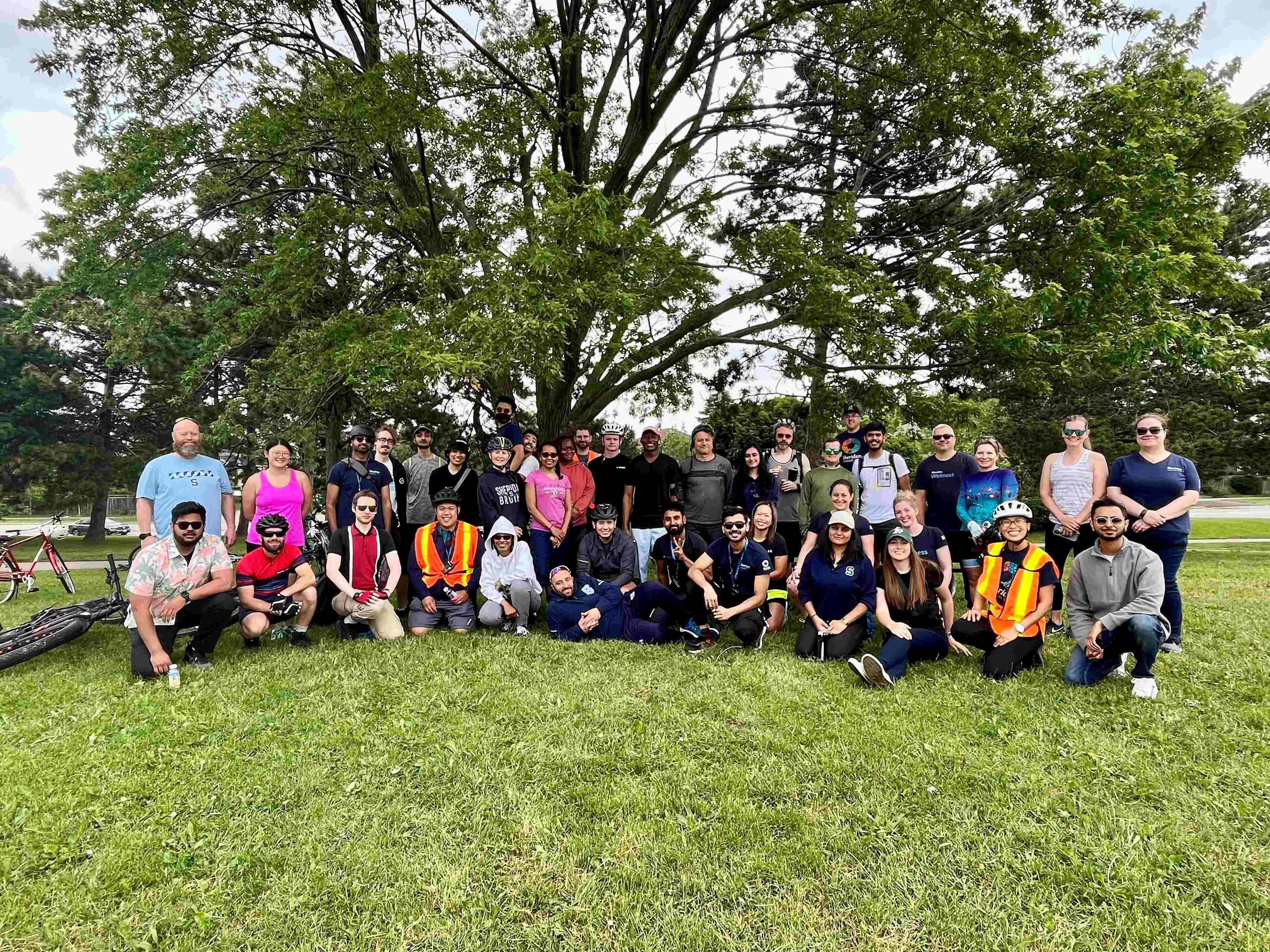 A group of Sheridan students and staff wearing cycling gear pose under a tree on the grass
