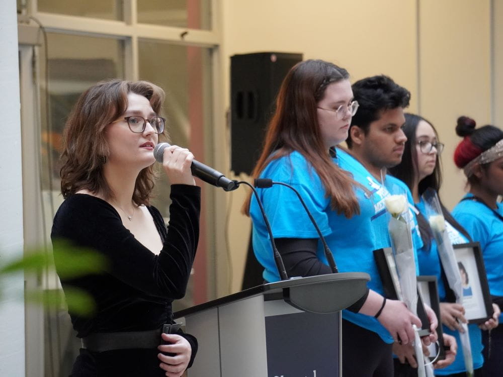 A student holds a microphone and sings. She is standing next to other students in blue shirts.