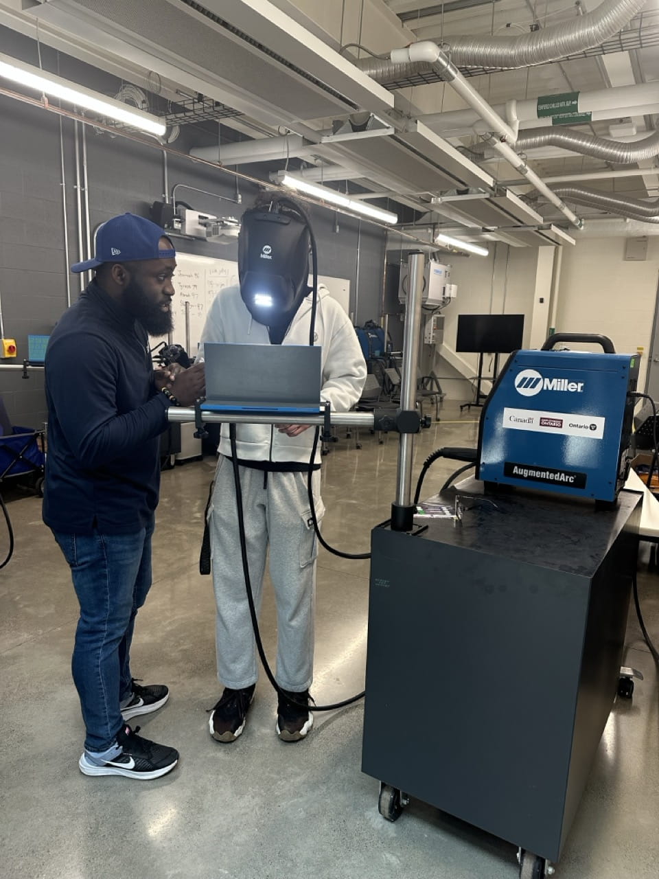A student wearing a welding mask receives instructions from another person