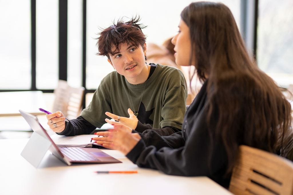 Two students in conversation at a table with a laptop