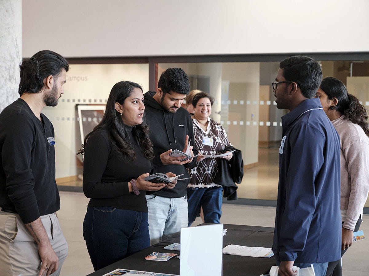 Two people talking to another across a table. Another person is observing the conversation while an additional person flips through a booklet.