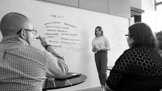 Person standing in front of whiteboard opposite two people seated