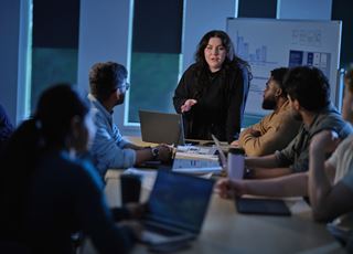 A student presenting to a group of her peers who are seated around a long table.