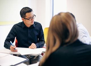 A student with paper and pen in hand, sitting at a table with two classmates