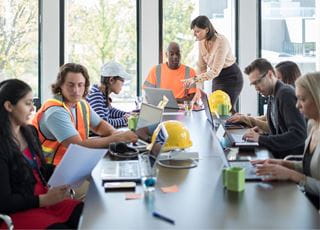 A group of students working on laptops around a long table, some wearing safety vests