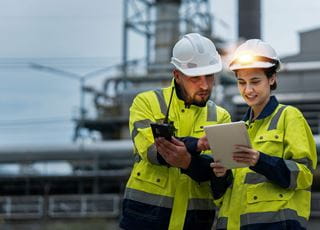Two people at a construction site wearing neon jackets and hard hats while looking at a tablet.