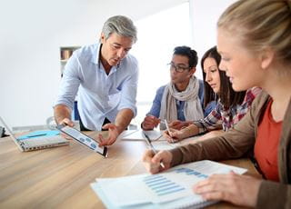 A man with grey hair showing three college students charts and graphs on a tablet while they sit at a table writing notes.