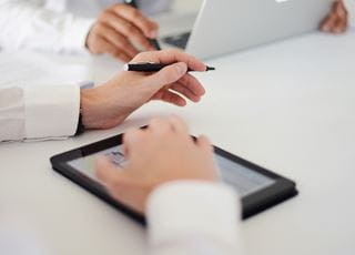Left hand holding a pen and right hand laid across a tablet.