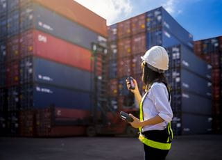 A young woman wearing a hard hat and fluorescent vest while standing near shipping containers