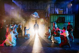 Group of actors on each side of the stage reach out toward a character standing at centre stage